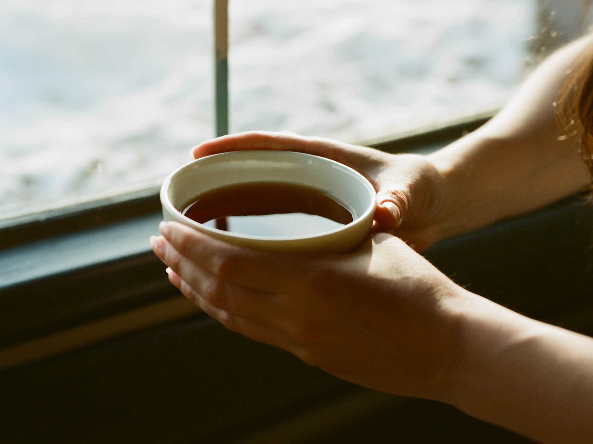 Manos de mujer sosteniendo una taza de té junto a ventana con luz invernal suave