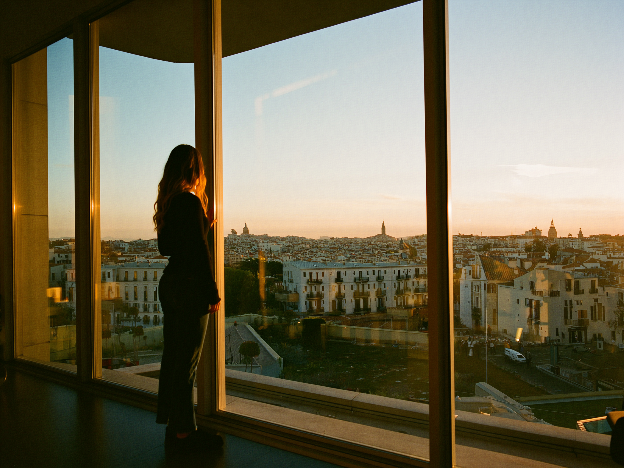 Mujer contemplando la ciudad desde su apartamento durante la hora dorada, transmitiendo paz y reflexión