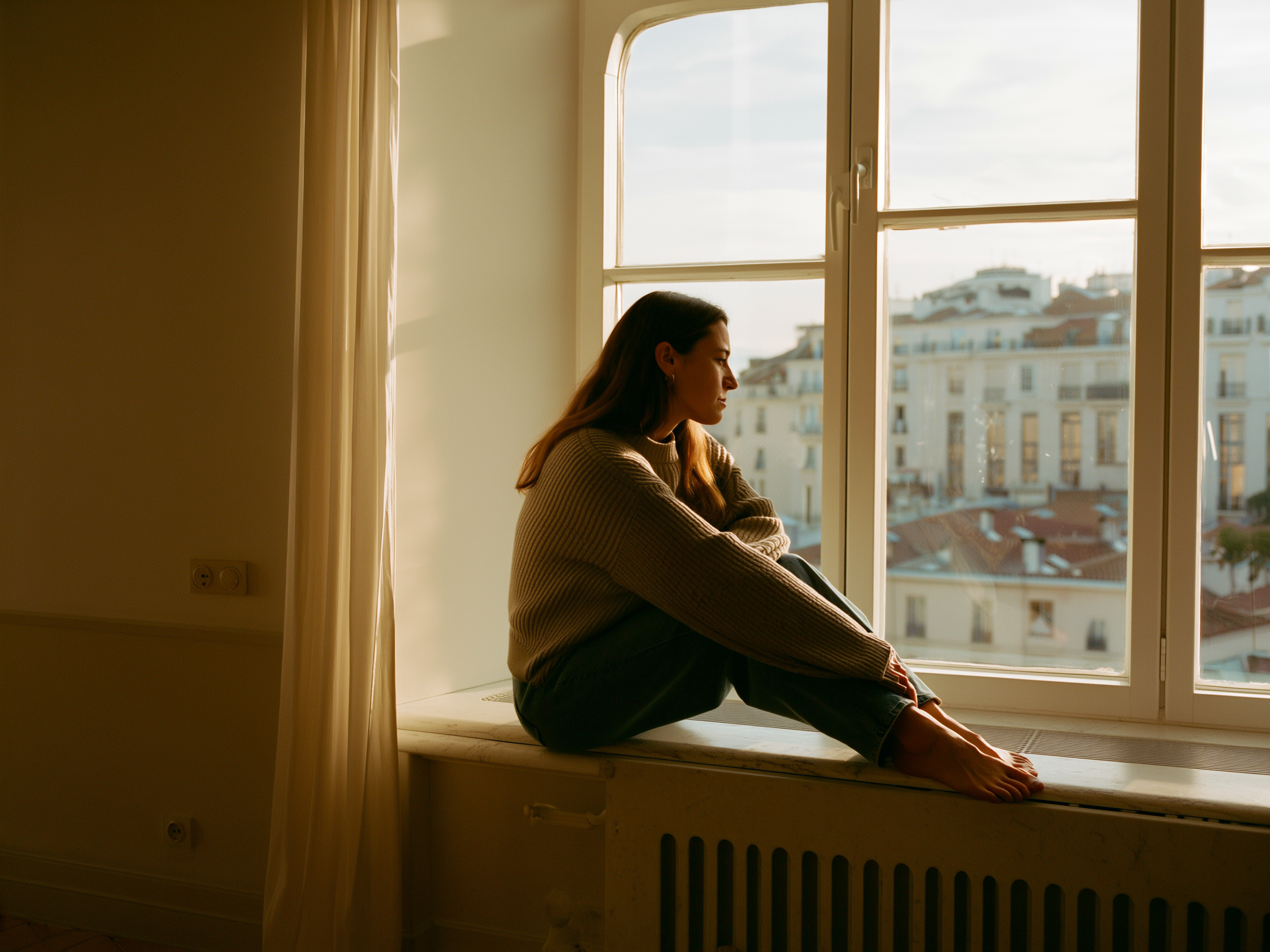 Mujer joven mirando por la ventana con expresión serena y esperanzadora en un ambiente cálido y luminoso