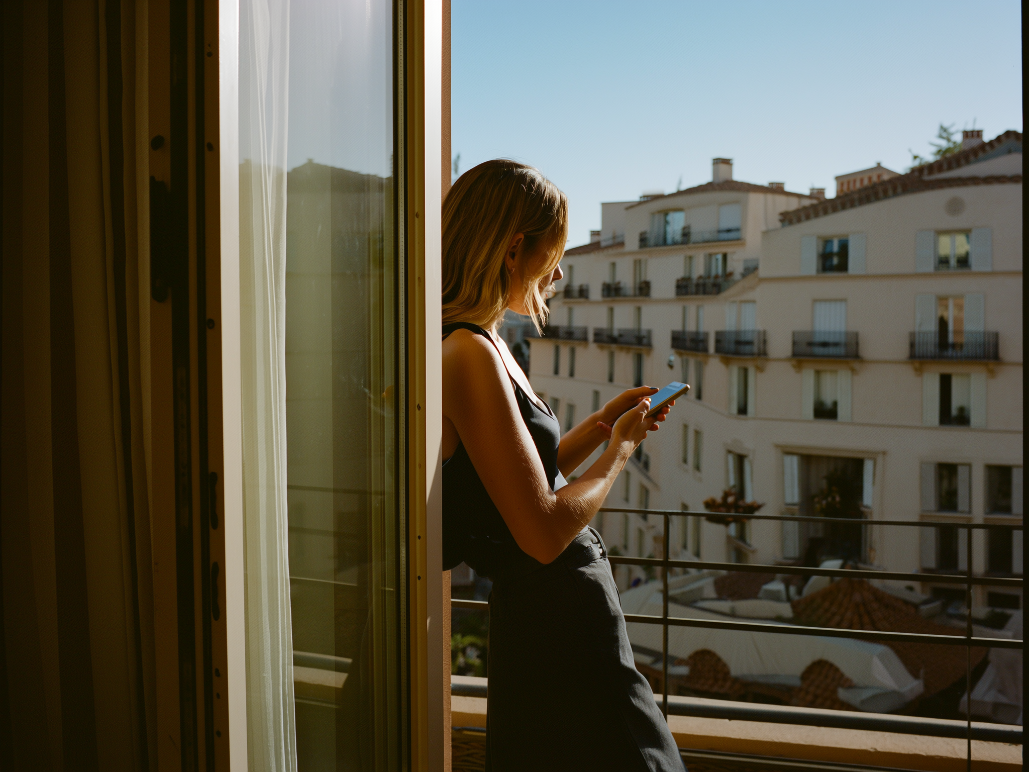 Mujer joven mirando pensativa por la ventana mientras sostiene su teléfono móvil, luz natural suave