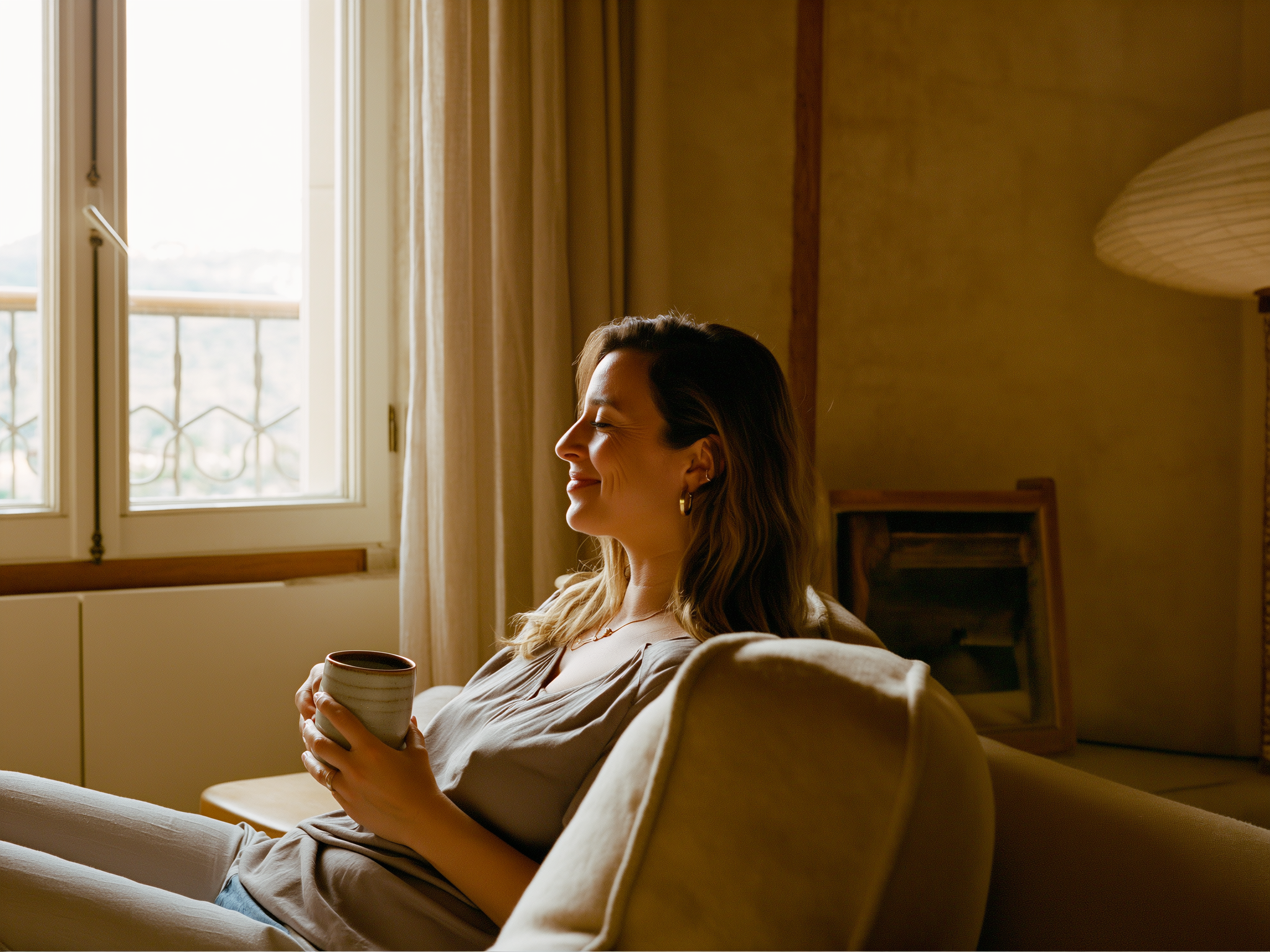 Mujer joven relajada en un sofá cómodo con una taza de té, mirando por la ventana con expresión serena